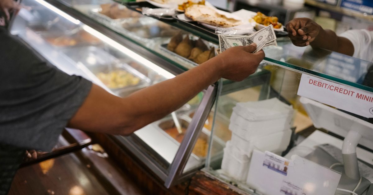 Crop faceless person standing near checkout buying food and passes money for dinner in cafe